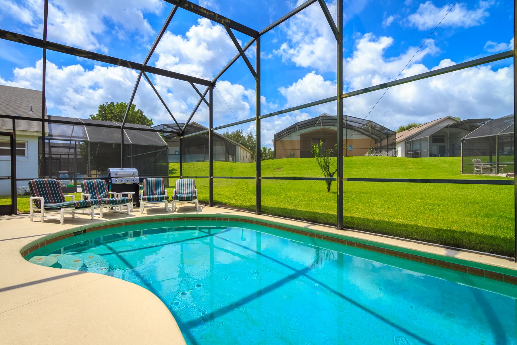 Screened lanai with a blue swimming pool and lounge chairs looking out on a green backyard